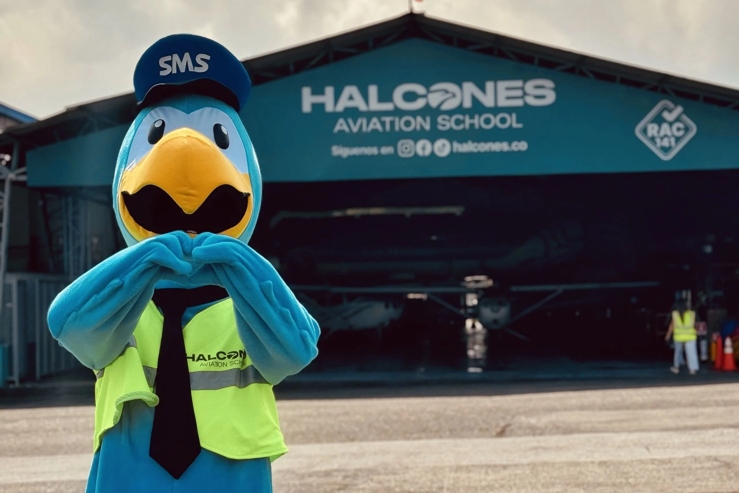 Mascota de Halcones Aviation School representando el Sistema de Gestión de Seguridad Operacional SMS frente a hangar aeronáutico.